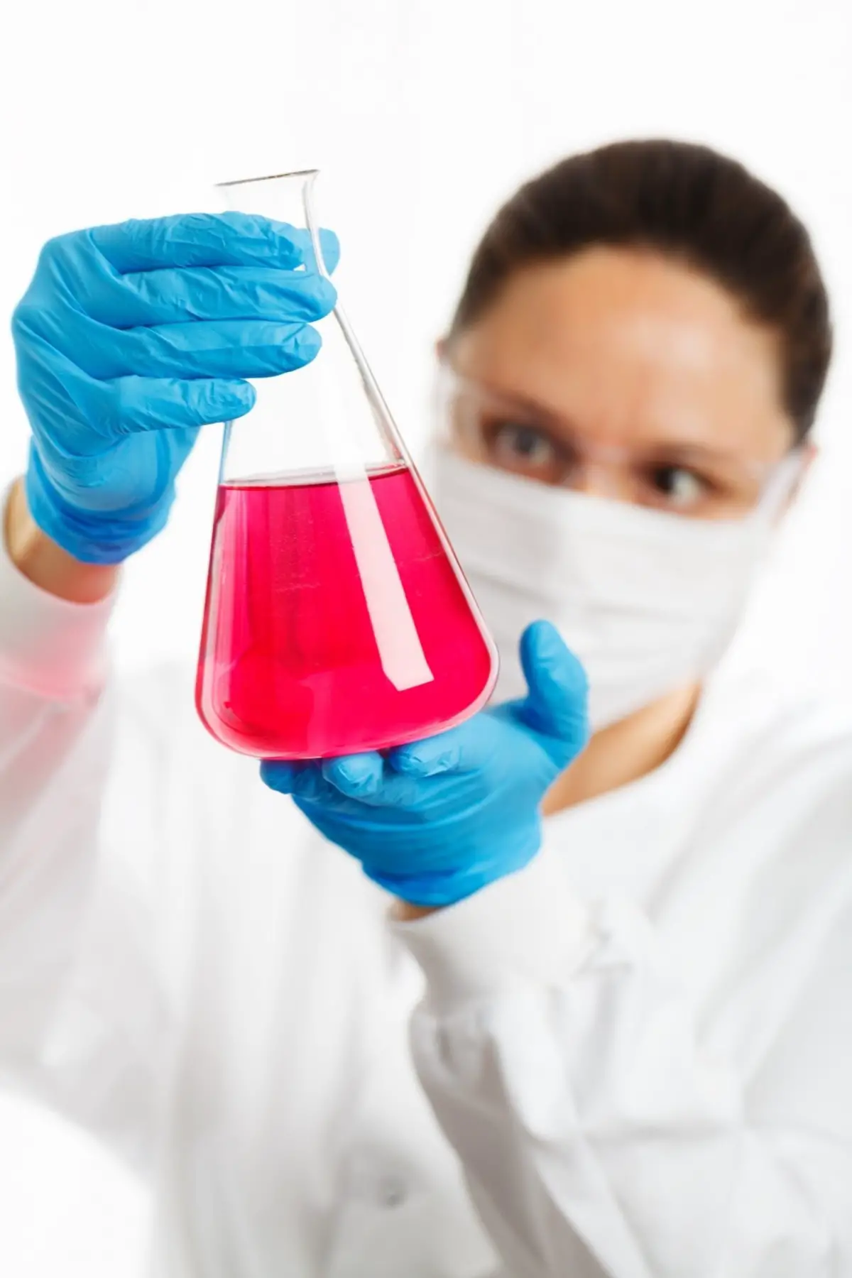 A picture of a researcher in a white cloak, blue gloves and protective glasses, holding a glass with pink liquid.