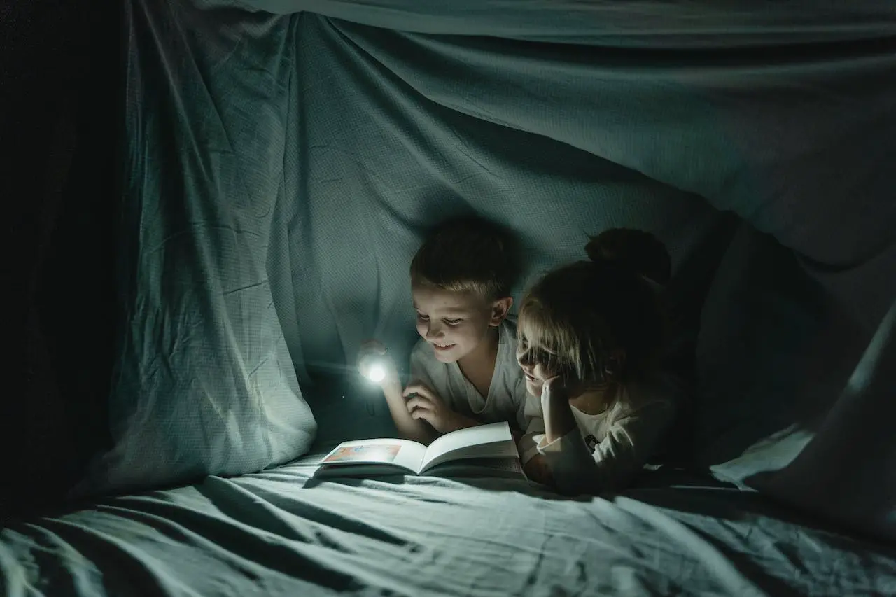 A picture of children reading a book witha flashlight inside a blanket fort.