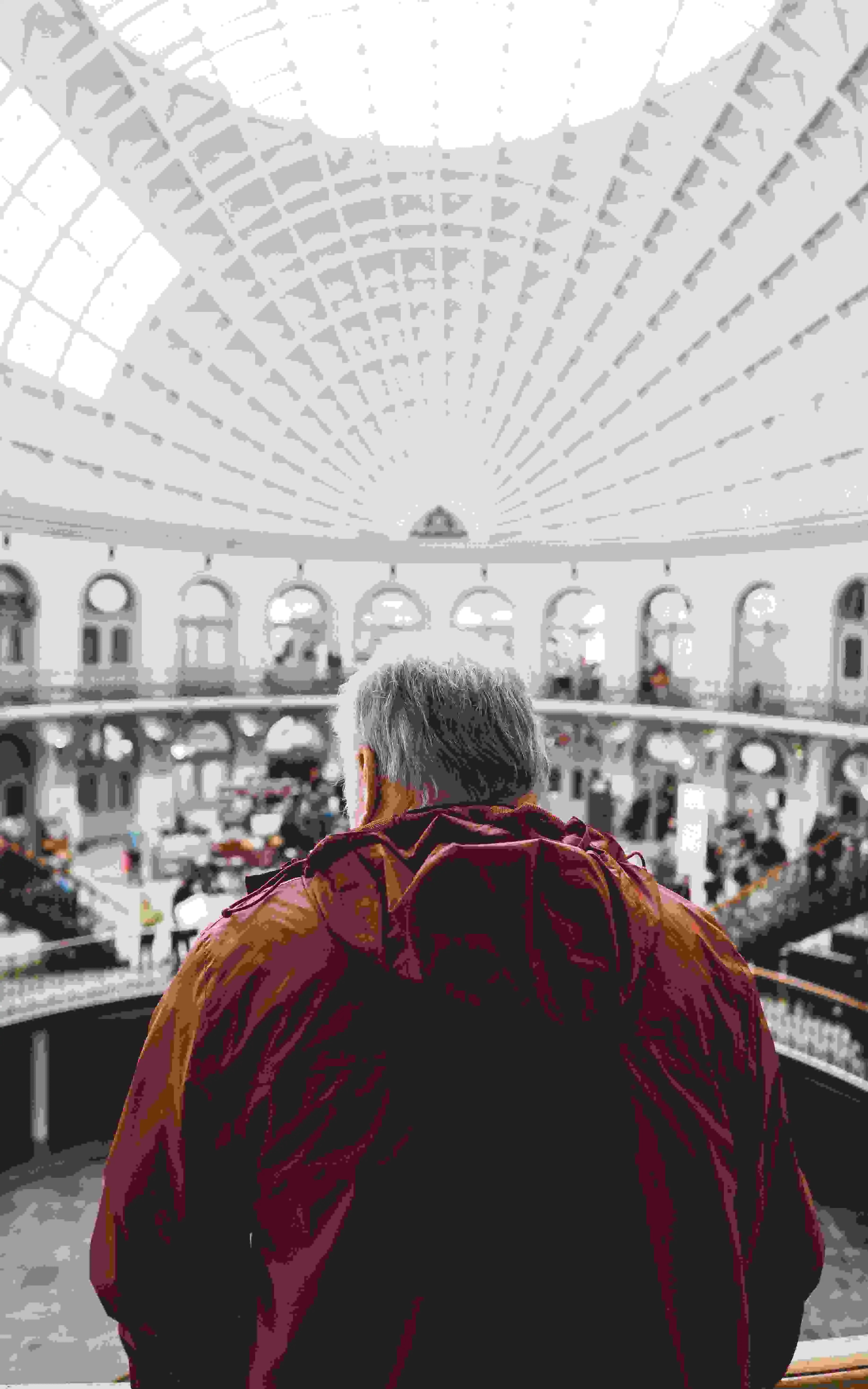 A older man with grey hair looking at the distance inside the museum, big rounded roof and arched windows evenly spaced out.