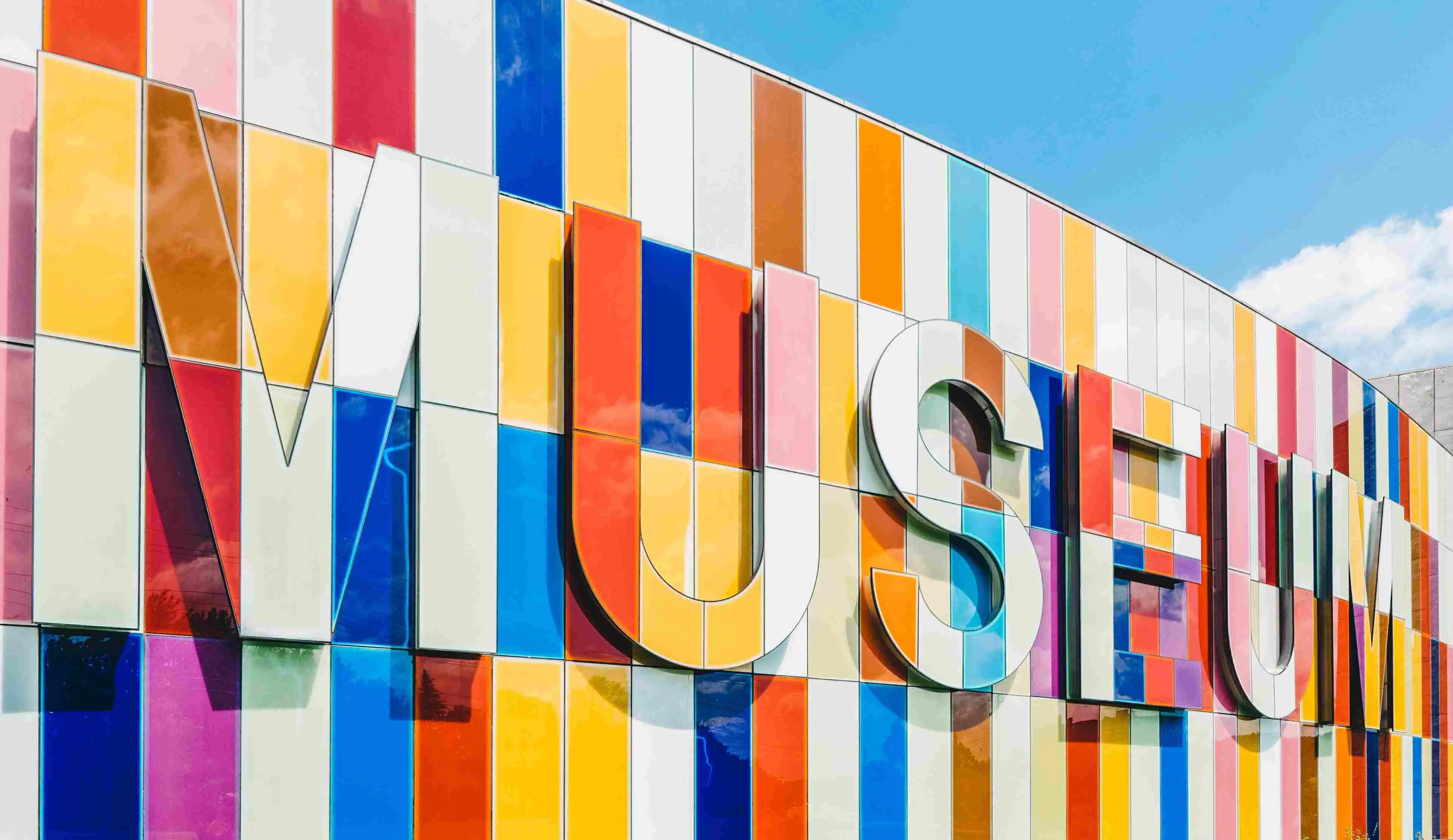 A picture take from the outside of the Musem, with the letters m, u, s, e, u, m physically sticking out of the building, and the whole building wall, including the letters, have been made of rectangular colorful tiles.