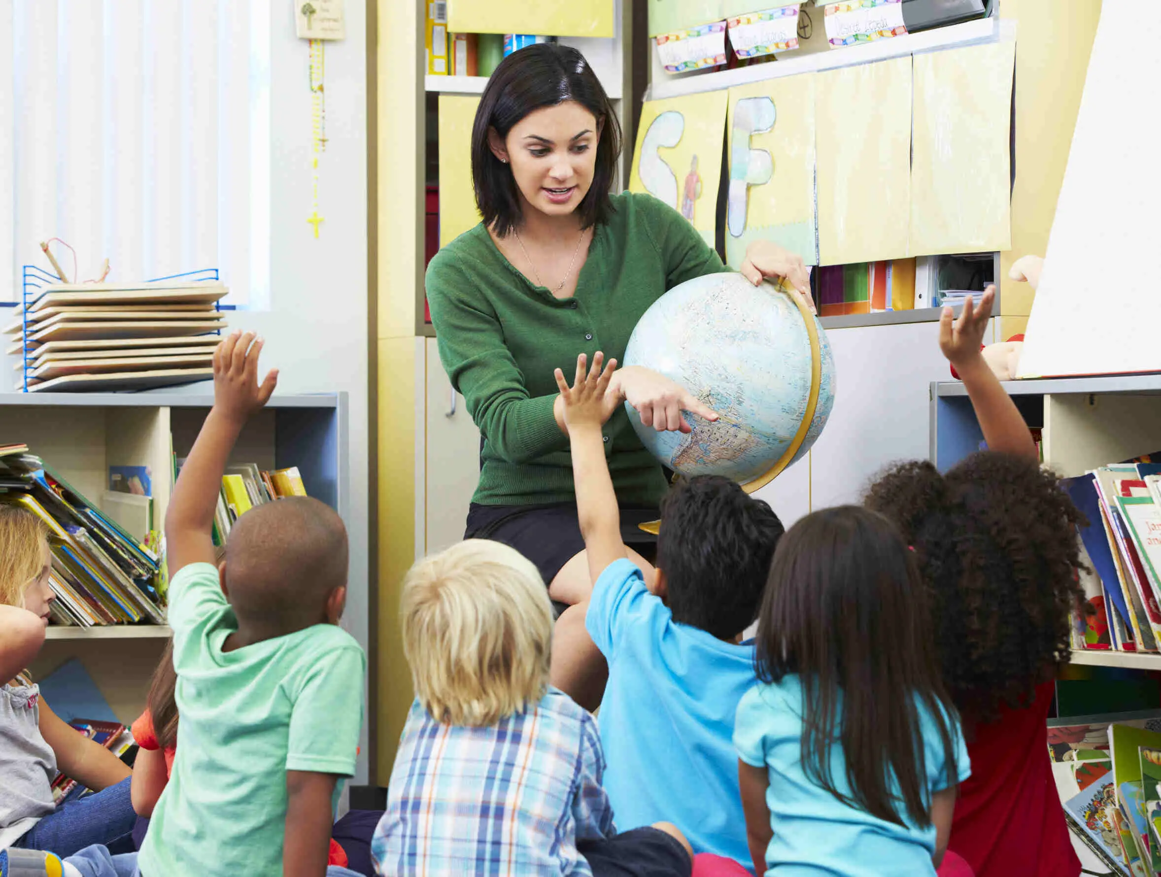 A picture of a young woman holding a globe and pointing at eager kids surrounding her.